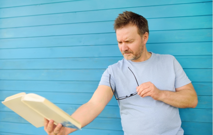 A man standing in front of a light blue wall, holding the book he is reading as far away as possible as he suffers from presbyopia