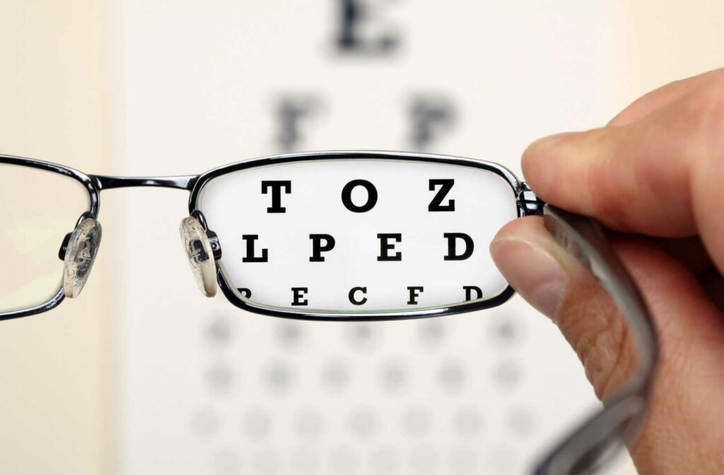 An eye chart can be seen clearly through the lens of a pair of glasses being held at the side by a hand.