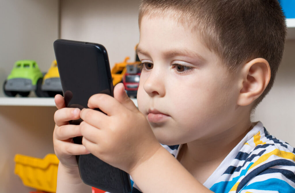 A small boy looks into the phone screen so close to his eyes. Can be a sign he has developing myopia.