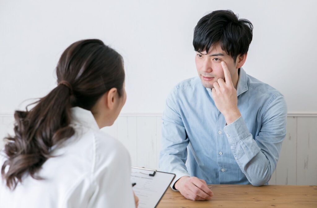 A man pointing to the corner of his left eye while talking to an attentive eye doctor.