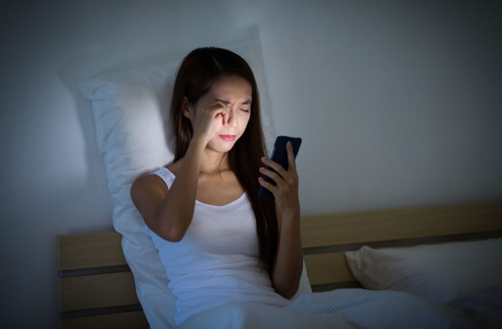 A young woman sitting on her bed in the dark before bed, looking at her cellphone, and rubbing her eyes.