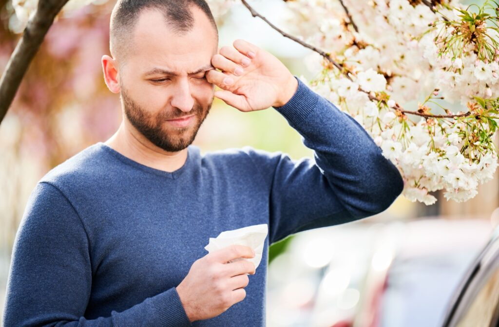 A young adult outdoors in springtime rubbing their eyes due to blurry vision during an allergy attack.