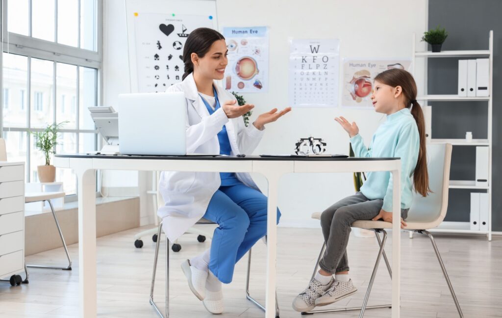 An optometrist sits chatting with a child in a brightly lit office after a comprehensive eye exam.