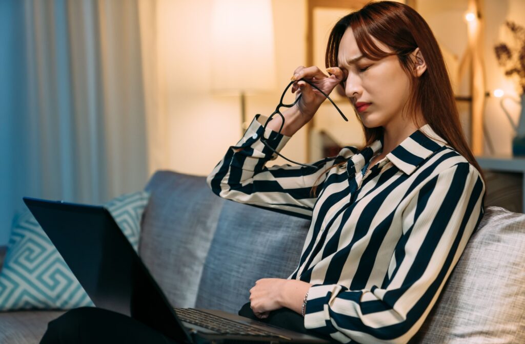 Woman in a striped shirt hold her glasses in her hands as she rubs her eyes while sitting in front of the computer