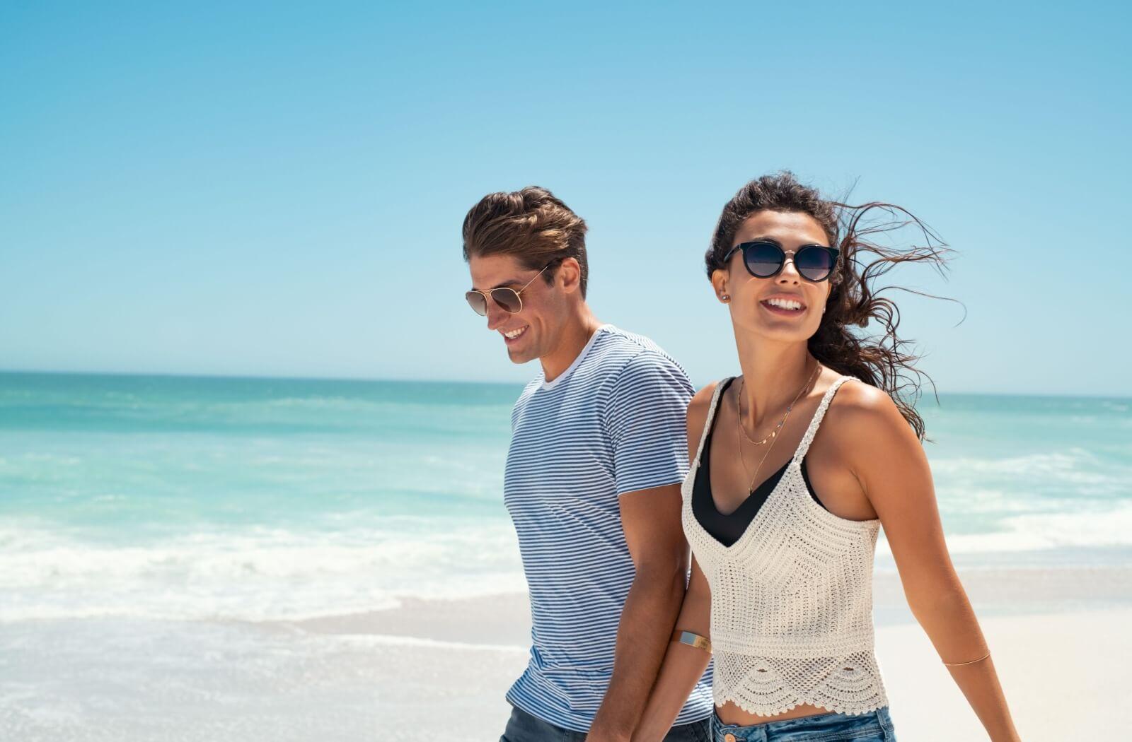 A smiling couple wearing polarized sunglasses enjoy a beach walk on a sunny day.
